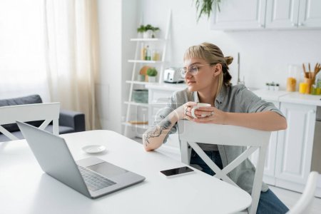 Foto de Mujer joven con tatuaje en la mano y flequillo sosteniendo taza de café y mirando a la computadora portátil cerca de teléfono inteligente y platillo en la mesa blanca alrededor de sillas en la cocina moderna, freelancer, estilo de vida remoto - Imagen libre de derechos