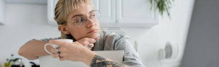 dreamy young woman with tattoo on hand and bangs holding cup of coffee and looking away near blurred laptop in modern kitchen, freelancer, remote lifestyle, eyeglasses, banner 