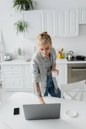 high anglve view of young woman with tattoo on hand and bangs holding cup of coffee and using laptop near smartphone and saucer on white table in modern kitchen, freelancer, remote lifestyle 