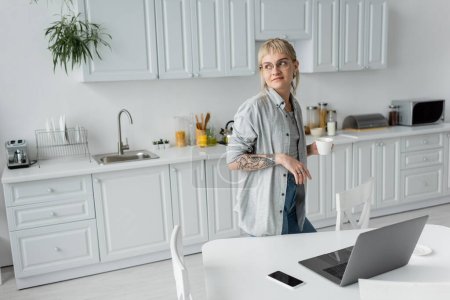 young woman with tattoo on hand and bangs holding cup of coffee and looking away while standing in modern kitchen next to laptop, smartphone with blank screen on white table near chairs, freelancer 