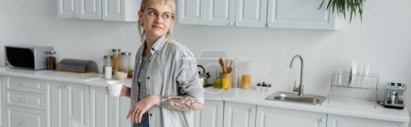 young woman in eyeglasses with tattoo on hand and bangs holding cup of coffee and looking away while standing in modern kitchen with different appliances and white cabinets, banner, morning time 