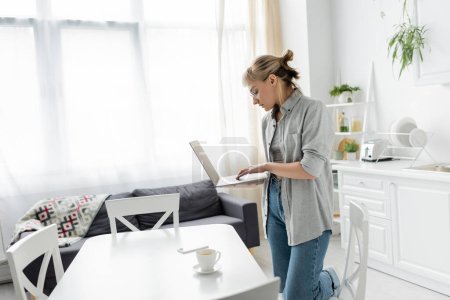 young woman in eyeglasses with short hair and bangs holding laptop near cup of coffee and smartphone on white table around chairs in white and modern kitchen next to sofa, freelancer, remote lifestyle
