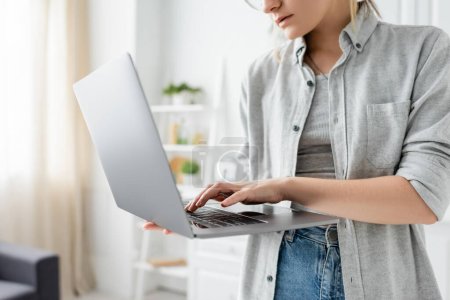 cropped view of focused young woman in grey shirt holding and using laptop in white and modern kitchen, blurred background, remote lifestyle, freelancer 