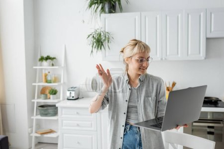 Photo for Happy young woman in eyeglasses, short hair and bangs holding laptop and waving hand during video call in white and modern kitchen, blurred background, remote lifestyle, freelancer - Royalty Free Image