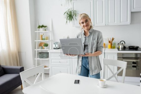 glückliche junge Frau in Brille mit kurzen Haaren und Pony hält Laptop in der Hand und blickt in die Kamera neben Tasse Kaffee und Smartphone mit leerem Bildschirm auf weißem Tisch in weißer und moderner Küche, freiberuflich