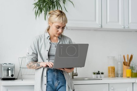 mujer joven tatuada en gafas, pelo corto y flequillo sosteniendo y utilizando el ordenador portátil mientras trabaja desde casa en la cocina blanca y moderna, fondo borroso, estilo de vida remoto, freelancer