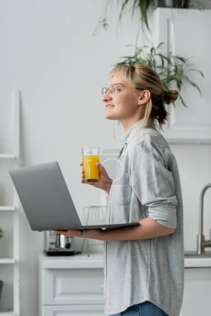 happy young woman in eyeglasses, short hair and bangs holding glass of fresh orange juice and laptop while working from home in white and modern kitchen, blurred background, freelancer