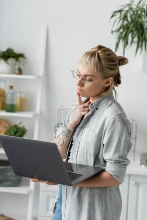 pensive young woman in eyeglasses, with tattoo and bangs using laptop while working from home in white and modern kitchen, blurred background, remote lifestyle, freelancer, hand near chin
