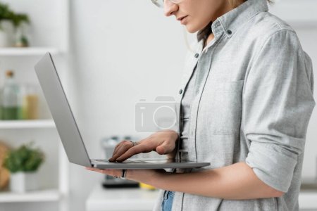 Foto de Vista recortada de la mujer joven enfocada en camisa gris sosteniendo y usando el ordenador portátil en la cocina blanca y moderna, fondo borroso, estilo de vida remoto, freelancer, trabajo desde casa, autónomo - Imagen libre de derechos