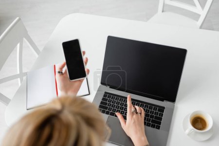 vista superior de la mujer borrosa sosteniendo teléfono inteligente con pantalla en blanco cerca de la computadora portátil, portátil con pluma, y una taza de café con platillo en la mesa blanca mientras trabaja desde casa, freelancer, espacio de trabajo moderno 