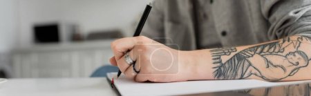 Photo for Cropped shot of young woman with tattoo on hand writing in notebook, taking notes, having inspiration while holding pen near laptop on white table, blurred foreground, work from home, banner - Royalty Free Image