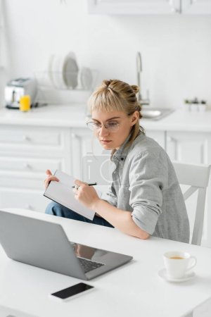 mujer joven con tatuaje en la mano y flequillo que sostiene el cuaderno, tomando notas cerca del teléfono inteligente y el ordenador portátil en la mesa blanca, fondo borroso, trabajo desde casa 