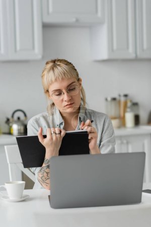 young woman in eyeglasses with tattoo on hand and bangs holding notebook, taking notes, sitting near laptop and cup of coffee on white table, blurred background, work from home 