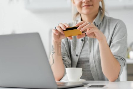 vista recortada de la joven feliz con tatuaje en la mano celebración de la tarjeta de crédito, sentado cerca de la computadora portátil, teléfono inteligente y taza de café en la mesa blanca, fondo borroso, trabajo desde casa 