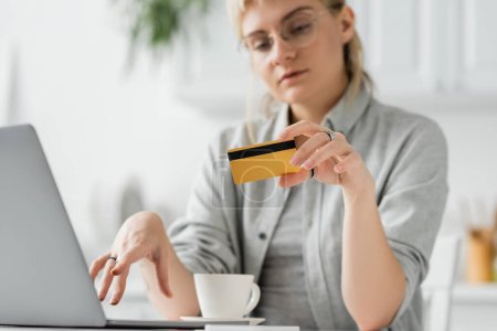 junge Frau mit Brille und tätowierter Kreditkarte in der Hand, neben Laptop und Kaffeetasse auf weißem Tisch sitzend, verschwommener Hintergrund, Arbeit von zu Hause aus, Online-Transaktionen, Technologie 