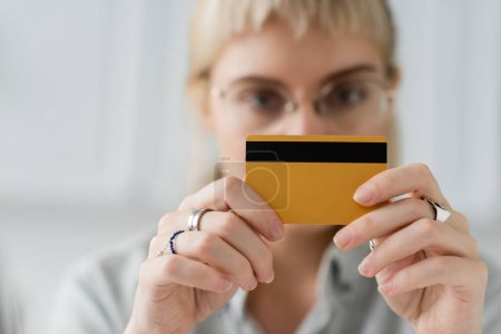 blurred scene of young woman in eyeglasses with rings on fingers holding credit card in hands and looking at camera at home with blurred background, copy space 