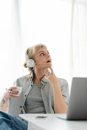 Photo for Pensive young woman with blonde hair, bangs and tattoo on hand sitting in wireless headphones and holding cup of coffee near laptop and blurred smartphone on table. freelance. work from home - Royalty Free Image