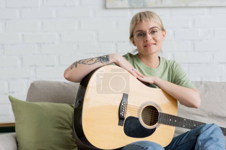 Téléchargez les photos : Heureux jeune femme dans des lunettes avec frange et tatouage à la main tenant la guitare acoustique et regardant la caméra tout en étant assis sur un canapé confortable dans le salon moderne à la maison - en image libre de droit