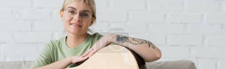 mujer joven satisfecha en gafas con flequillo y tatuaje en la mano sosteniendo la guitarra acústica y mirando a la cámara mientras está sentado en la sala de estar moderna en casa, sonrisa, pancarta 