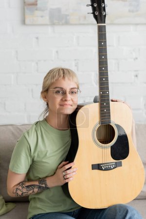mujer tatuada y feliz en gafas con flequillo sosteniendo la guitarra acústica y sentada en un cómodo sofá en la sala de estar moderna, aprendiendo música, desarrollo de habilidades, entusiasta de la música, mira a la cámara