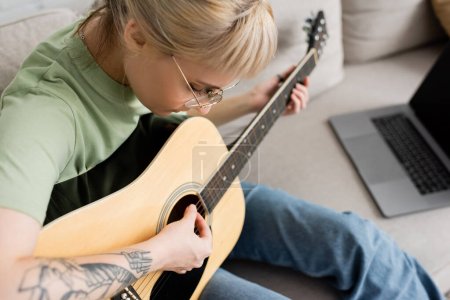 junge Frau in Brille mit Pony und Tätowierung, die eine akustische Gitarre in der Hand hält und lernt, wie man in der Nähe von Laptop spielt und auf einer bequemen Couch im modernen Wohnzimmer zu Hause sitzt, virtueller Unterricht