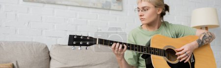 young woman in glasses with bangs and tattoo on hand playing acoustic guitar and sitting on comfortable couch in modern living room, learning music, skill development, music enthusiast, banner 