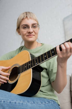 Photo for Happy young woman in glasses with bangs playing acoustic guitar and sitting on comfortable couch in modern living room, learning music, skill development, music enthusiast - Royalty Free Image