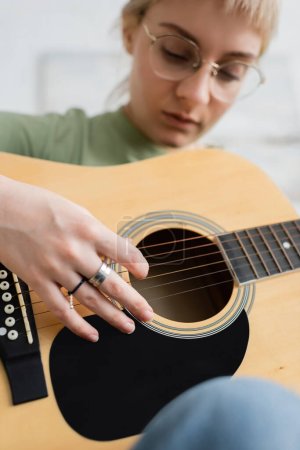 Foto de Joven talanted en gafas con flequillo, anillos en los dedos tocando la guitarra acústica y sentado en la sala de estar moderna, aprendiendo música, desarrollo de habilidades, entusiasta de la música - Imagen libre de derechos
