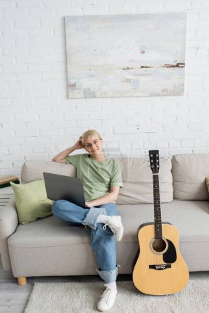 young woman with blonde and short hair, bangs and eyeglasses using laptop while sitting on comfortable couch and looking at camera near guitar in modern living room with painting on wall 