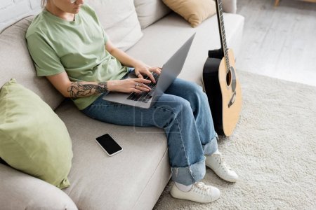vista recortada de la mujer joven con tatuaje en la mano usando el ordenador portátil mientras está sentado en un cómodo sofá al lado del teléfono inteligente y la guitarra en la sala de estar moderna, freelance, trabajo desde casa