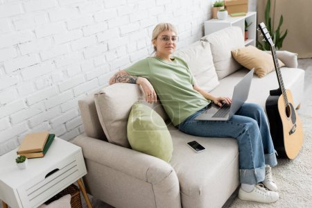 mujer tatuada feliz con el pelo rubio y corto, flequillo y gafas usando el ordenador portátil mientras está sentado en un cómodo sofá al lado del teléfono inteligente y mirando a la cámara cerca de la guitarra en la sala de estar moderna 