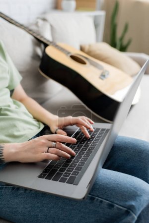 Ausgeschnittene Ansicht einer verschwommenen jungen Freiberuflerin mit Tätowierung an der Hand mittels Laptop, während sie im modernen Wohnzimmer auf einer bequemen Couch neben der Gitarre sitzt, freiberuflich, von zu Hause aus arbeitend