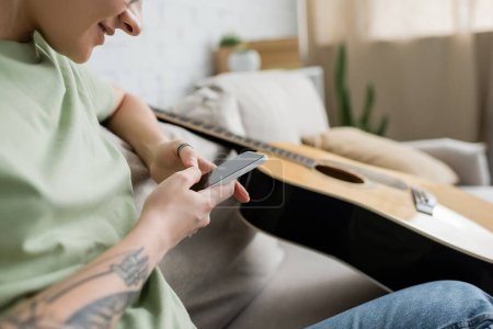 cropped view of happy young woman with tattoo on hand using smartphone while sitting on comfortable couch near guitar in modern living room, blurred shot 