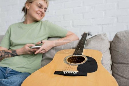 glückliche junge Frau mit blonden und kurzen Haaren, Pony und Brille mit Smartphone, während sie im modernen Wohnzimmer auf einer bequemen Couch neben der Gitarre sitzt, unscharfe Aufnahme 