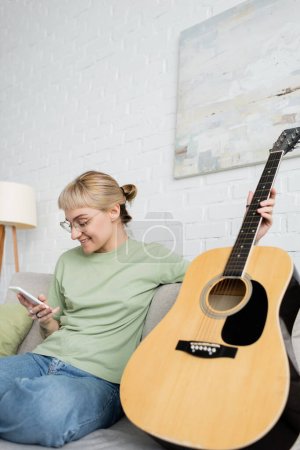 happy young woman with blonde and short hair, bangs and eyeglasses using smartphone while sitting on comfortable couch near guitar in modern living room with painting on wall 