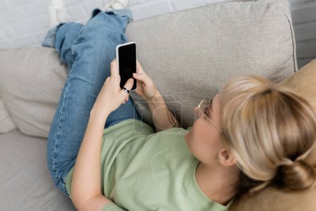 Foto de Mujer joven con cabello rubio y corto y anteojos, tatuaje en la mano y ropa casual usando teléfono inteligente mientras descansa en un cómodo sofá, vaqueros, camiseta - Imagen libre de derechos