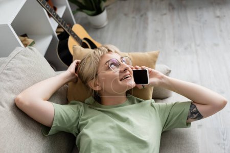 overhead view of happy young woman with blonde and short hair, bangs and eyeglasses talking on smartphone while resting on comfortable couch near guitar in modern living room 