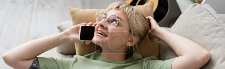 overhead view of happy young woman with blonde and short hair, bangs and eyeglasses talking on smartphone while resting on comfortable couch near guitar in modern living room, banner 