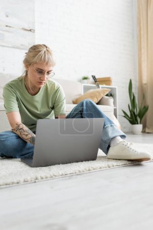 mujer tatuada con cabello rubio, flequillo y anteojos usando computadora portátil mientras está sentada en una alfombra cerca de un cómodo sofá, planta borrosa y estante en la moderna sala de estar con paiting en la pared 