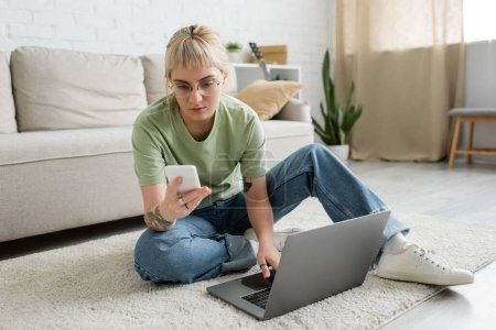 tattooed woman with bangs and eyeglasses using laptop while sitting on carpet and holding smartphone near comfortable couch and rack with plants in modern living room 