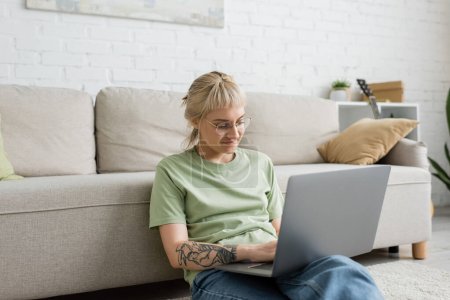 tattooed woman with blonde and short hair, bangs and eyeglasses typing on laptop while sitting on carpet near comfortable couch in modern living room with paiting on wall 