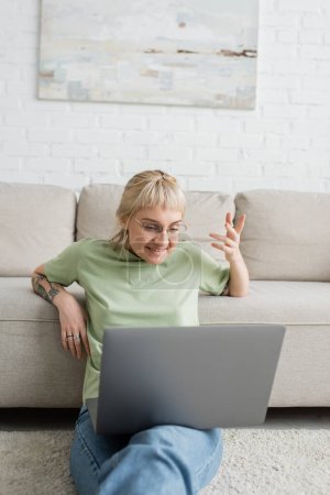 Tätowierte Frau mit blonden und kurzen Haaren, Pony und Brille, die beim Videoanruf auf dem Laptop lächelt, während sie auf einem Teppich in der Nähe einer bequemen Couch im modernen Wohnzimmer mit Wandmalerei sitzt 