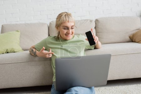 cheerful and tattooed woman with bangs and eyeglasses using laptop while sitting on carpet and holding smartphone with blank screen near comfortable couch in modern living room 