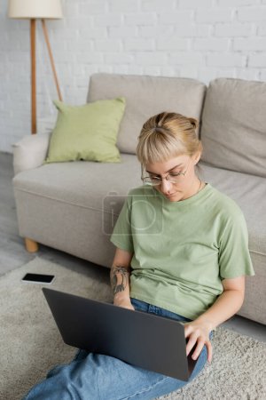 Téléchargez les photos : Femme tatouée avec une frange et des lunettes à l'aide d'un ordinateur portable tout en étant assis sur le tapis près du smartphone avec écran blanc et canapé confortable dans le salon moderne - en image libre de droit