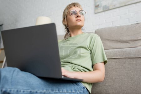 low angle view of pensive woman with blonde and short hair, bangs and eyeglasses using laptop while sitting near comfortable couch in modern living room with paiting on wall 