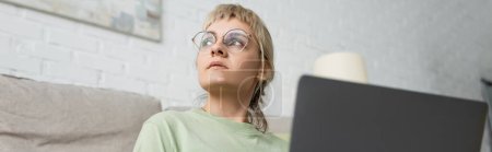 Photo for Low angle view of pensive woman with blonde and short hair, bangs and eyeglasses using laptop while sitting near comfortable couch in modern living room with paiting on wall, banner - Royalty Free Image