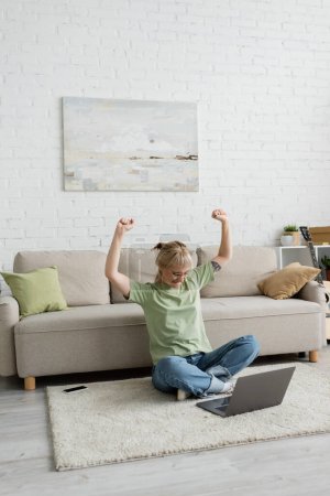excited tattooed woman in eyeglasses using laptop while sitting with raised hands on carpet near smartphone, comfortable couch, guitar and rack with plants in modern living room with paiting on wall 