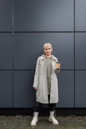 Photo for Stylish young woman with blonde hair with bangs standing in coat and hoodie while holding paper cup with coffee and looking at camera near grey modern building on street, outside, urban living - Royalty Free Image