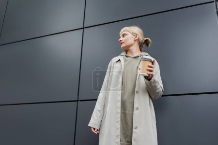 low angle view of stylish young woman with blonde hair with bangs standing in coat and hoodie while holding paper cup with takeaway coffee near grey modern building on street, outside, urban living 