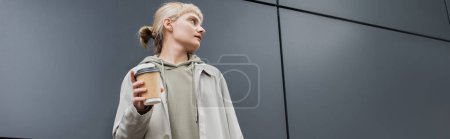Foto de Mujer joven de moda con cabello rubio con flequillo de pie en abrigo y sudadera con capucha mientras sostiene la taza de papel con café para llevar cerca de edificio moderno gris en la calle, exterior, vida urbana, pancarta - Imagen libre de derechos